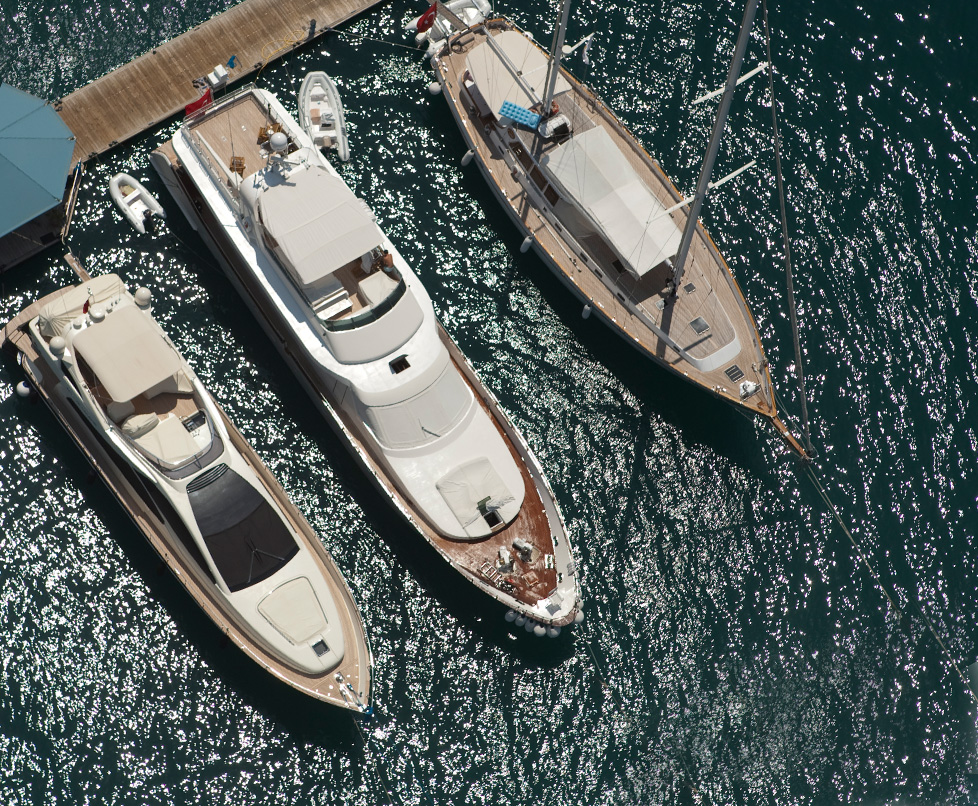 Mehrere Yachten liegen nebeneinander an einem Pier in einem ruhigen Hafen, das Wasser spiegelt den blauen Himmel.